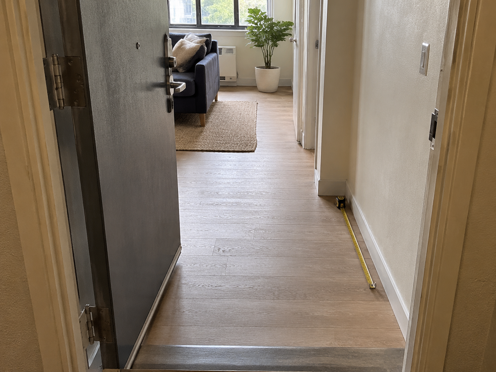 An open apartment door frames a clear entry path and standing area, with a tape measure marking the narrow wall-side zone left after door movement is accounted for.