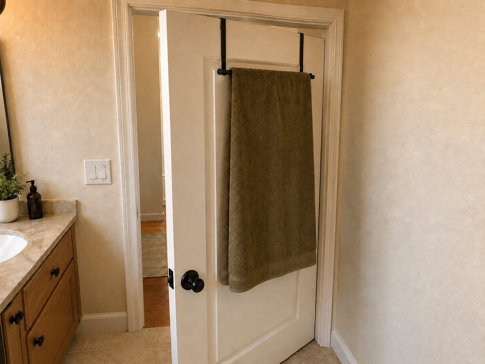 A bath towel hangs open from a slim over-door bar on a partly open bathroom door, clearing the adjacent wall, vanity, toilet, and floor.