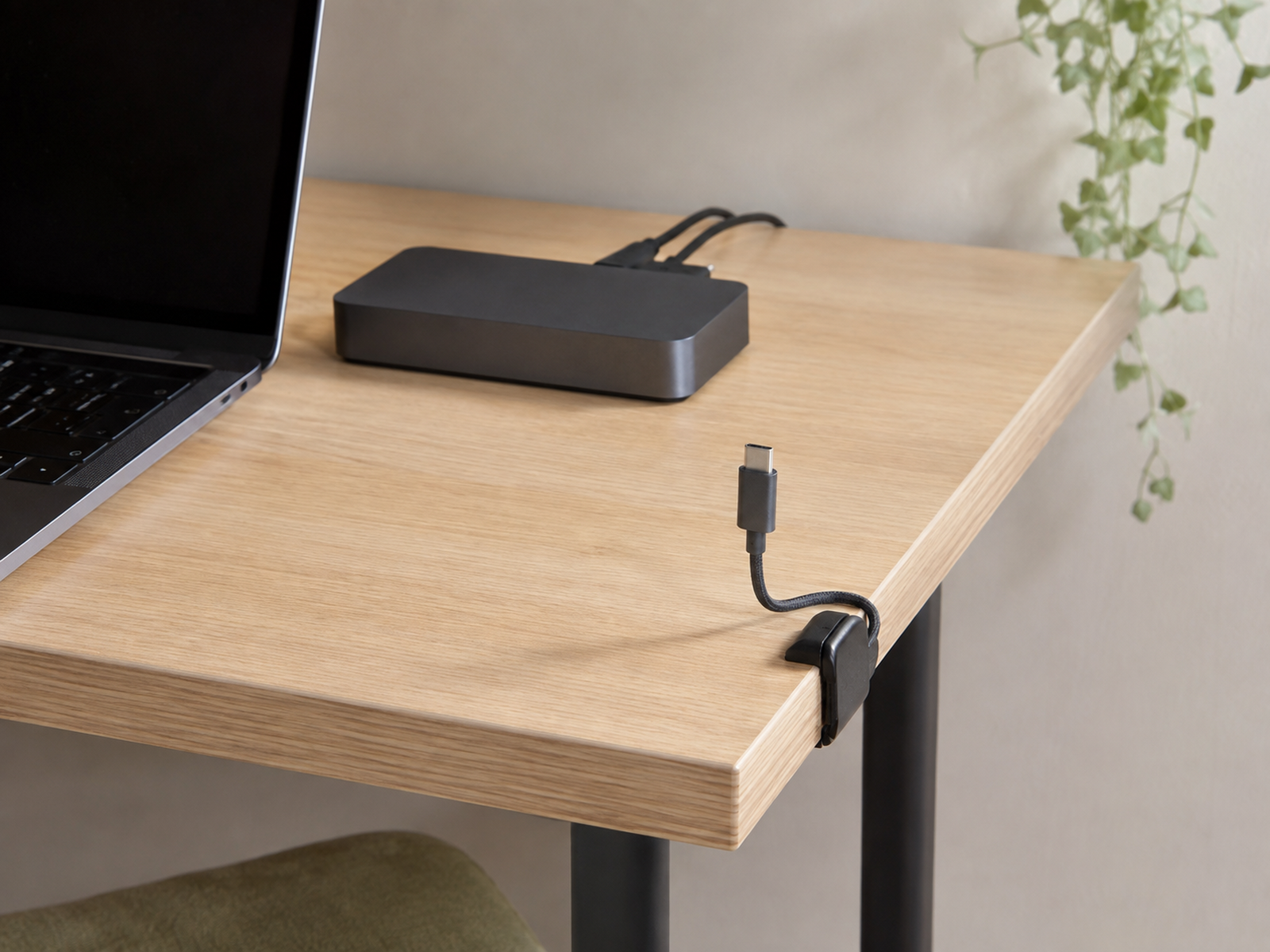 A USB-C charging cable is held upright at the edge of a small desk beside a reachable dock with fixed cables routed away.