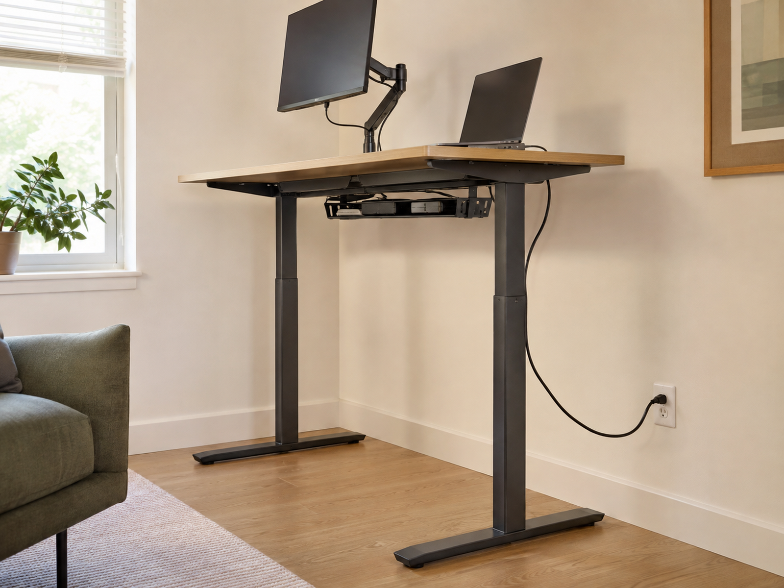 A raised sit-stand desk carries its power strip under the desktop while one relaxed cable descends beside the leg to a wall outlet.