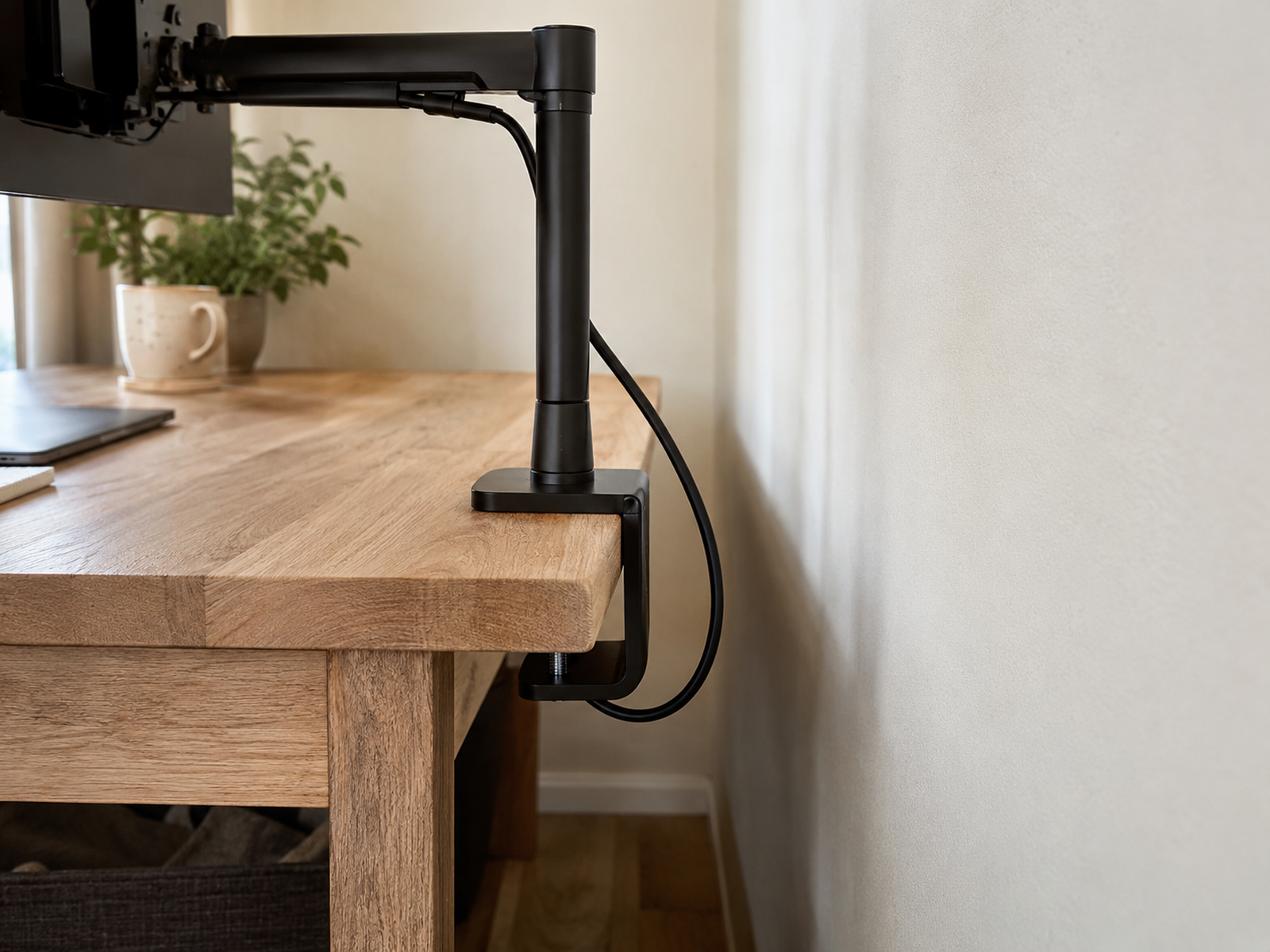 A close view shows a monitor-arm clamp fully gripping the rear edge of a shallow wood desk, with open underside access, a small wall gap, and a loose cable loop.