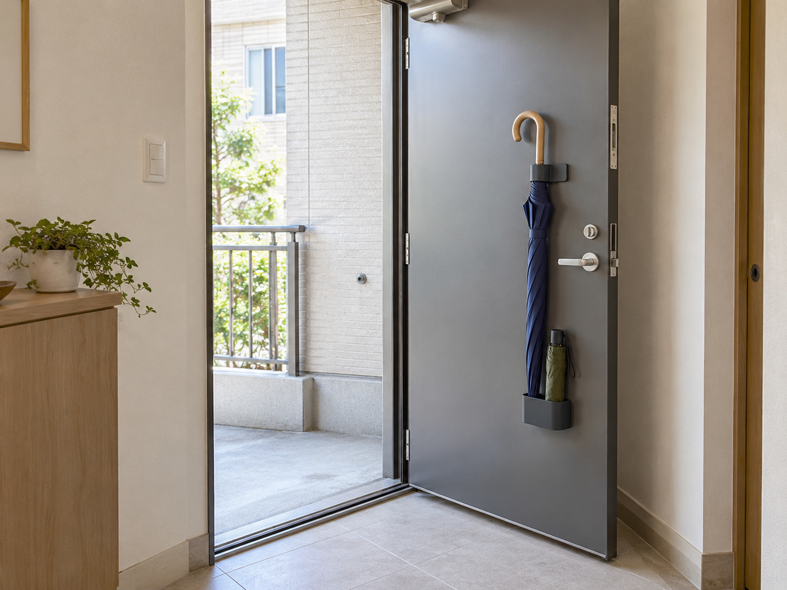 A dry long umbrella and folding umbrella stored on magnetic supports attached to an open steel apartment door, with the handle, threshold, and floor clear.