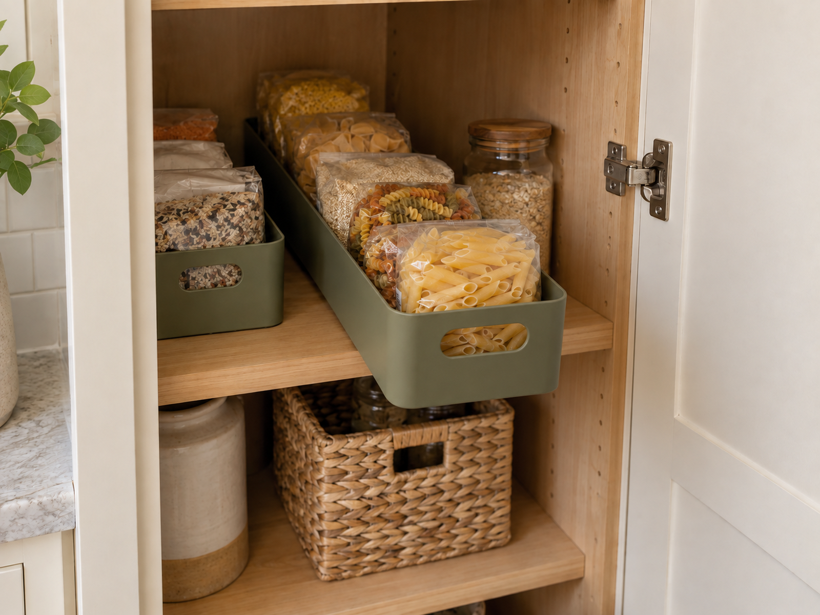 A long olive pantry bin partially pulled from a deep shelf, revealing pasta and grain packets from front to back while clearing the cabinet hinge.
