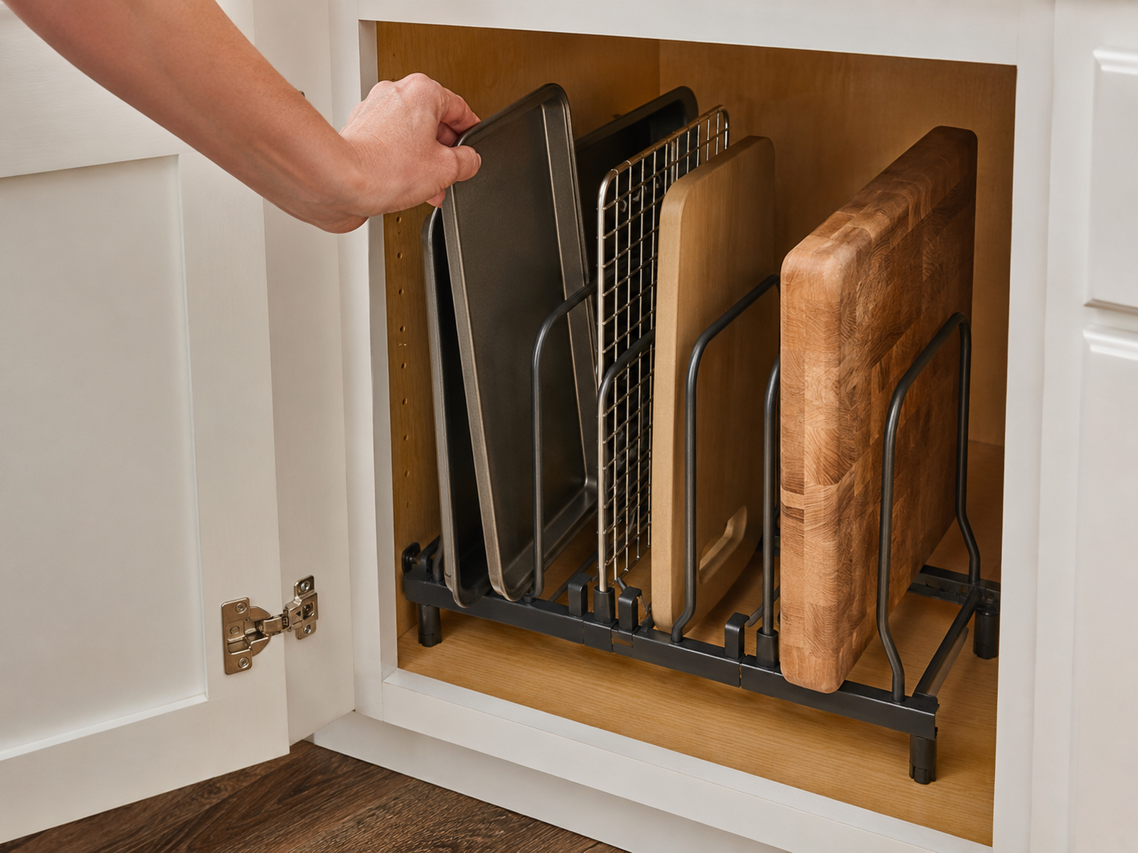 A hand lifts one baking sheet from a freestanding cabinet rack while thin pans and a cooling rack remain in narrow slots beside a wide slot holding a thick wood cutting board.